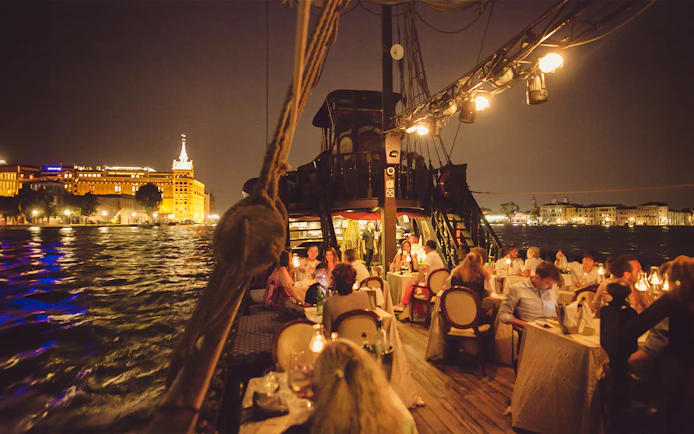 Dinner guests on a galleon cruise in Venice with city lights reflecting on the water.