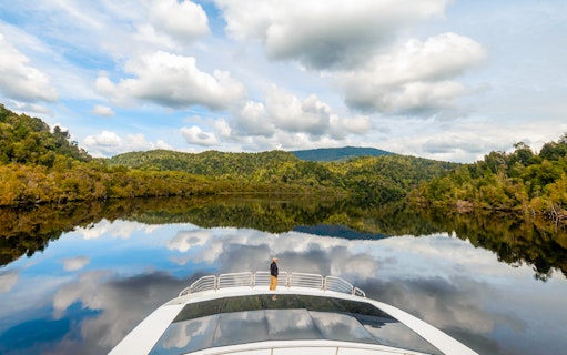 Man on cruise boat on Gordon River, surrounded by lush forest and calm water reflections.