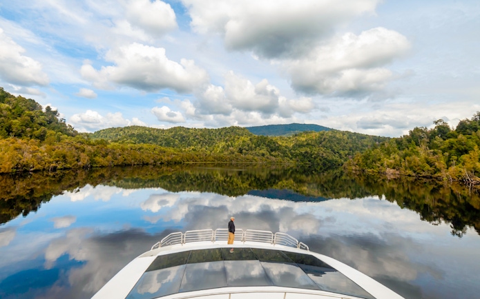 Man on cruise boat on Gordon River, surrounded by lush forest and calm water reflections.