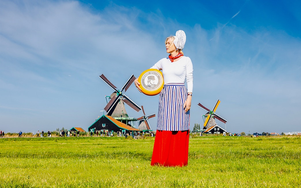 Person holding cheese wheel at Zaanse Schans with windmills in the background.