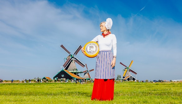 Person holding cheese wheel at Zaanse Schans with windmills in the background.