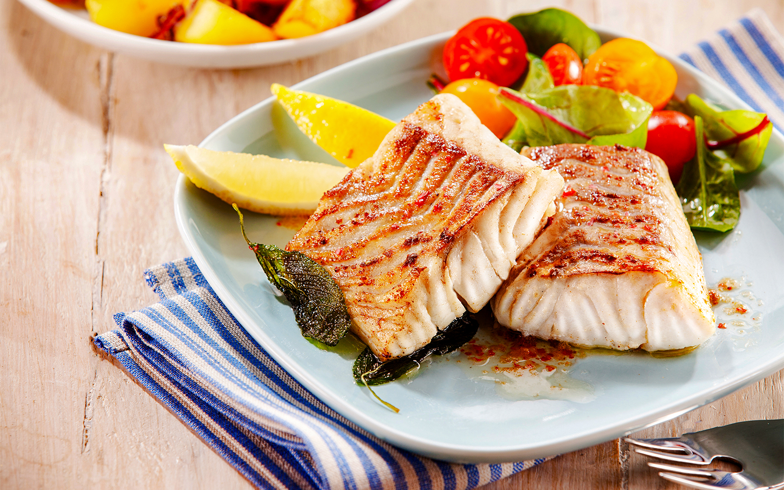 Grilled pollock fillets with lemon wedges and salad on a plate.