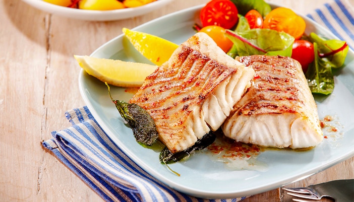 Grilled pollock fillets with lemon wedges and salad on a plate.