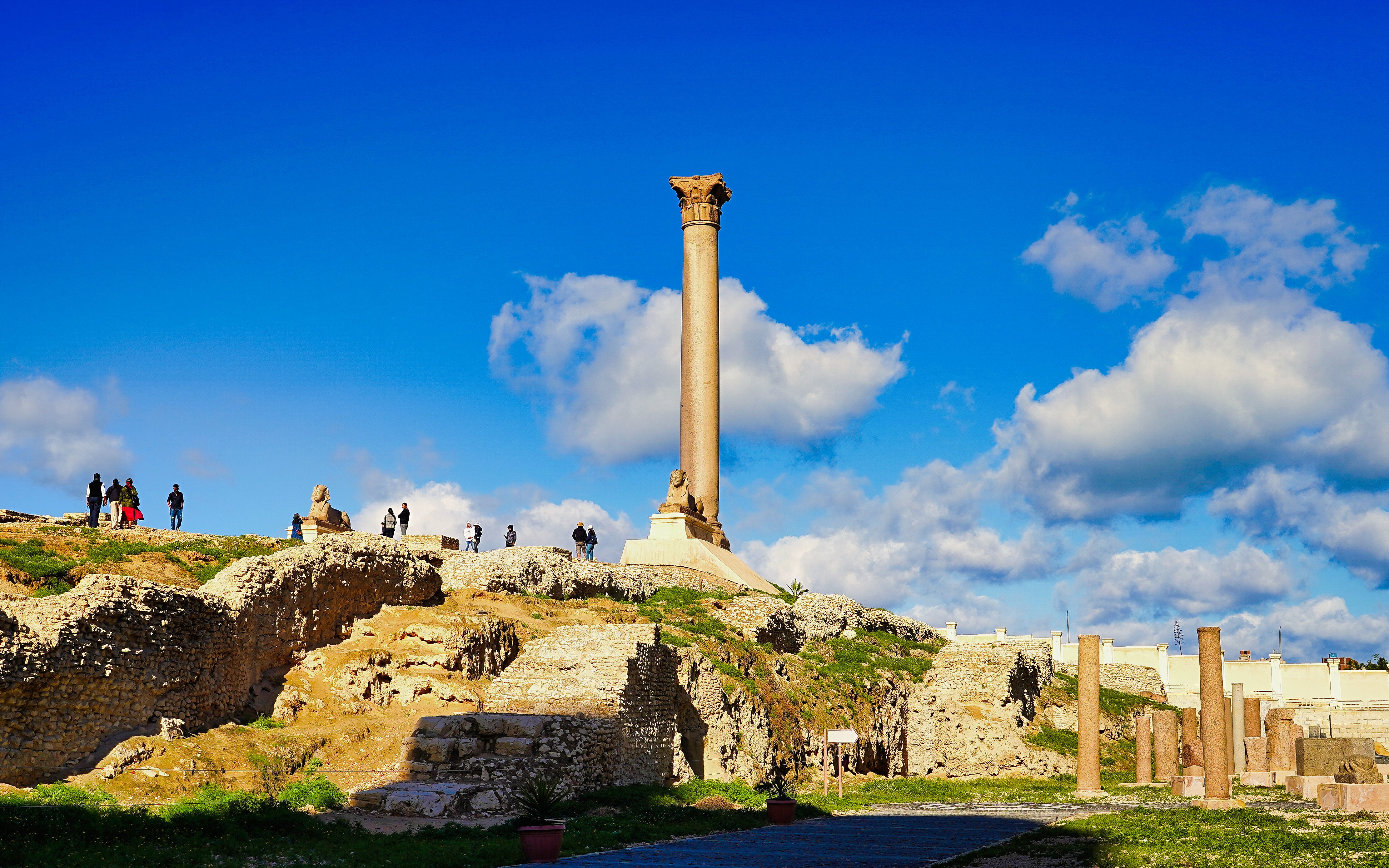 Pompey's Pillar in Alexandria, Egypt, with tourists exploring the ancient ruins.