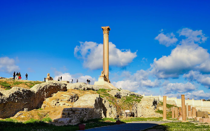 Pompey's Pillar in Alexandria, Egypt, with tourists exploring the ancient ruins.