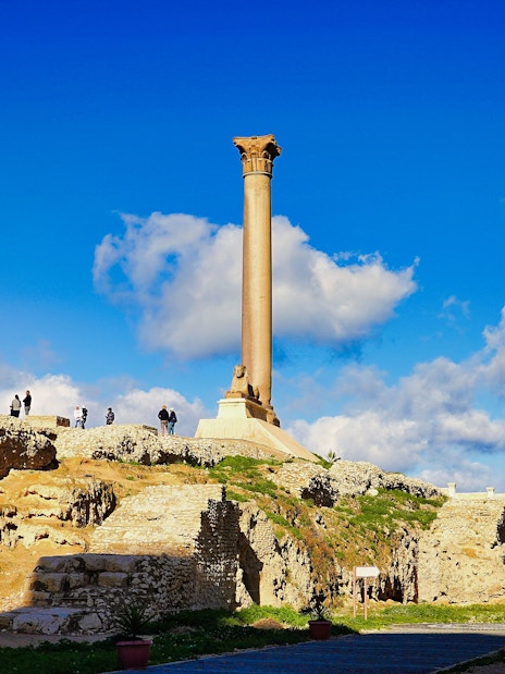 Pompey's Pillar in Alexandria, Egypt, with tourists exploring the ancient ruins.