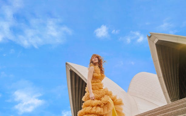 Person in a flowing dress on steps of Sydney Opera House under blue sky.
