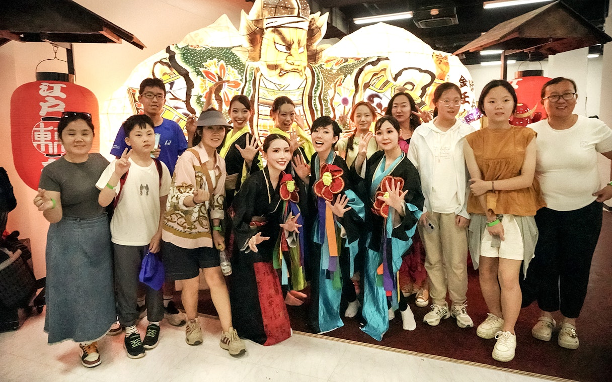 Dancers in traditional attire posing with tourists in front of a vibrant festival lantern.
