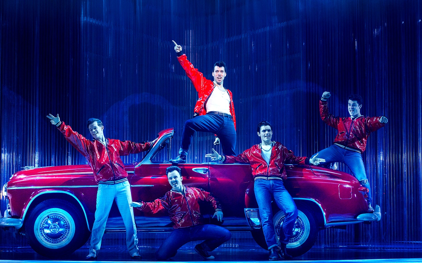 Performers in red jackets pose on stage with a vintage car during Grease in Brisbane.