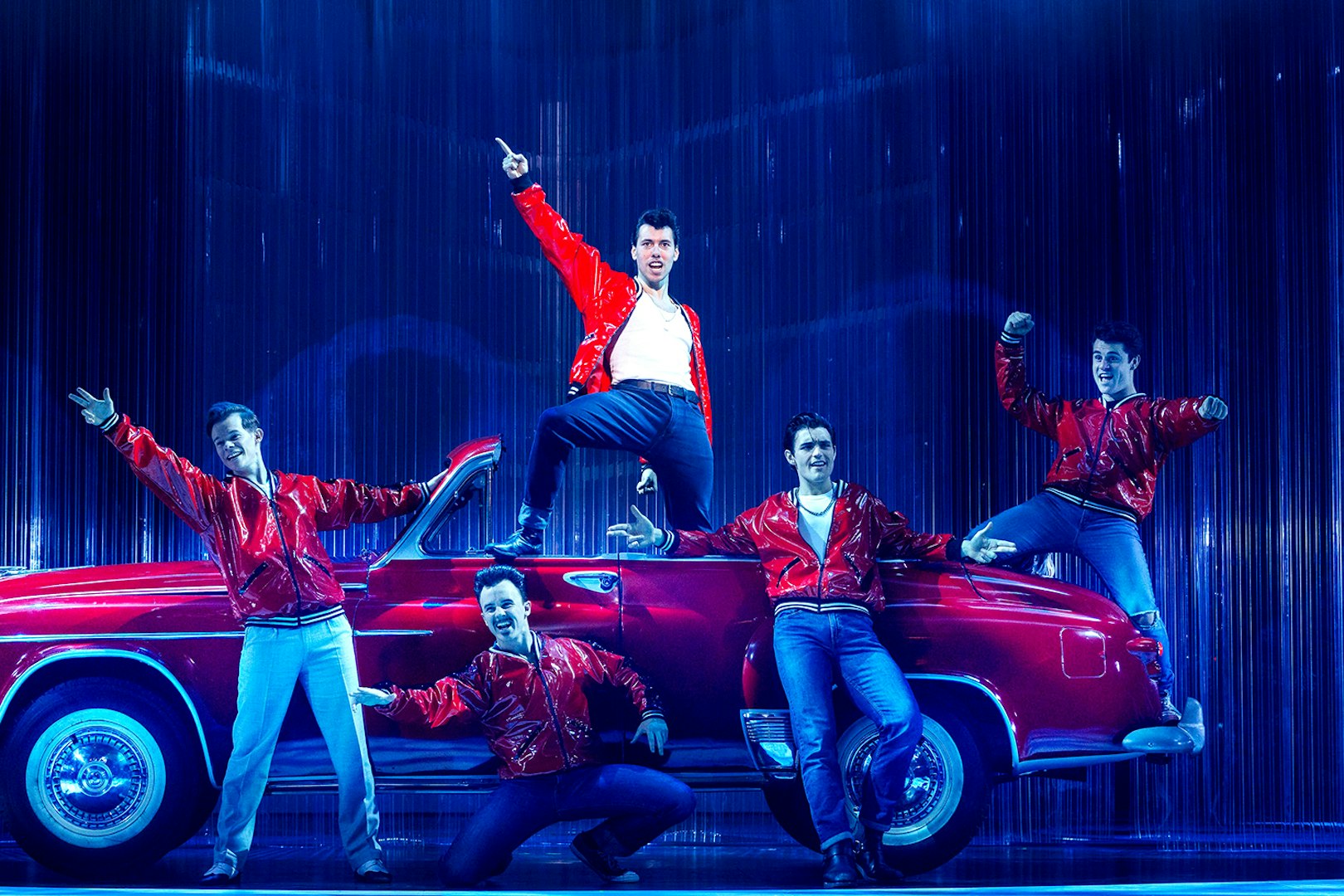 Performers in red jackets pose on stage with a vintage car during Grease in Brisbane.