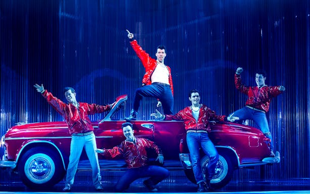 Performers in red jackets pose on stage with a vintage car during Grease in Brisbane.