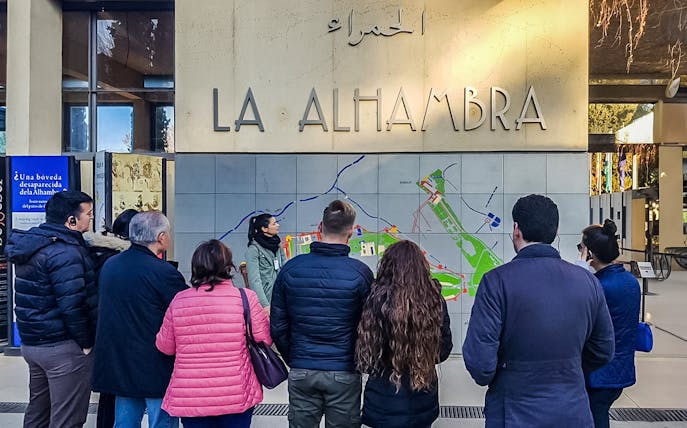 Visitors gather at Alhambra entrance for a guided tour, viewing a large map.