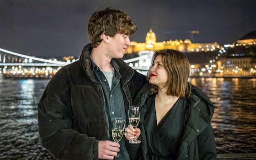 Guests with drinks on Danube sightseeing cruise, Budapest night skyline in background.