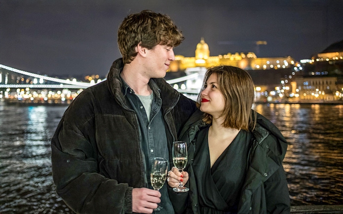 Guests with drinks on Danube sightseeing cruise, Budapest night skyline in background.