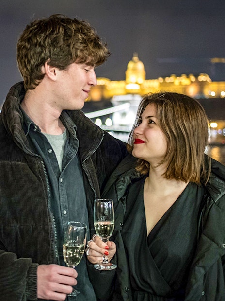 Guests with drinks on Danube sightseeing cruise, Budapest night skyline in background.