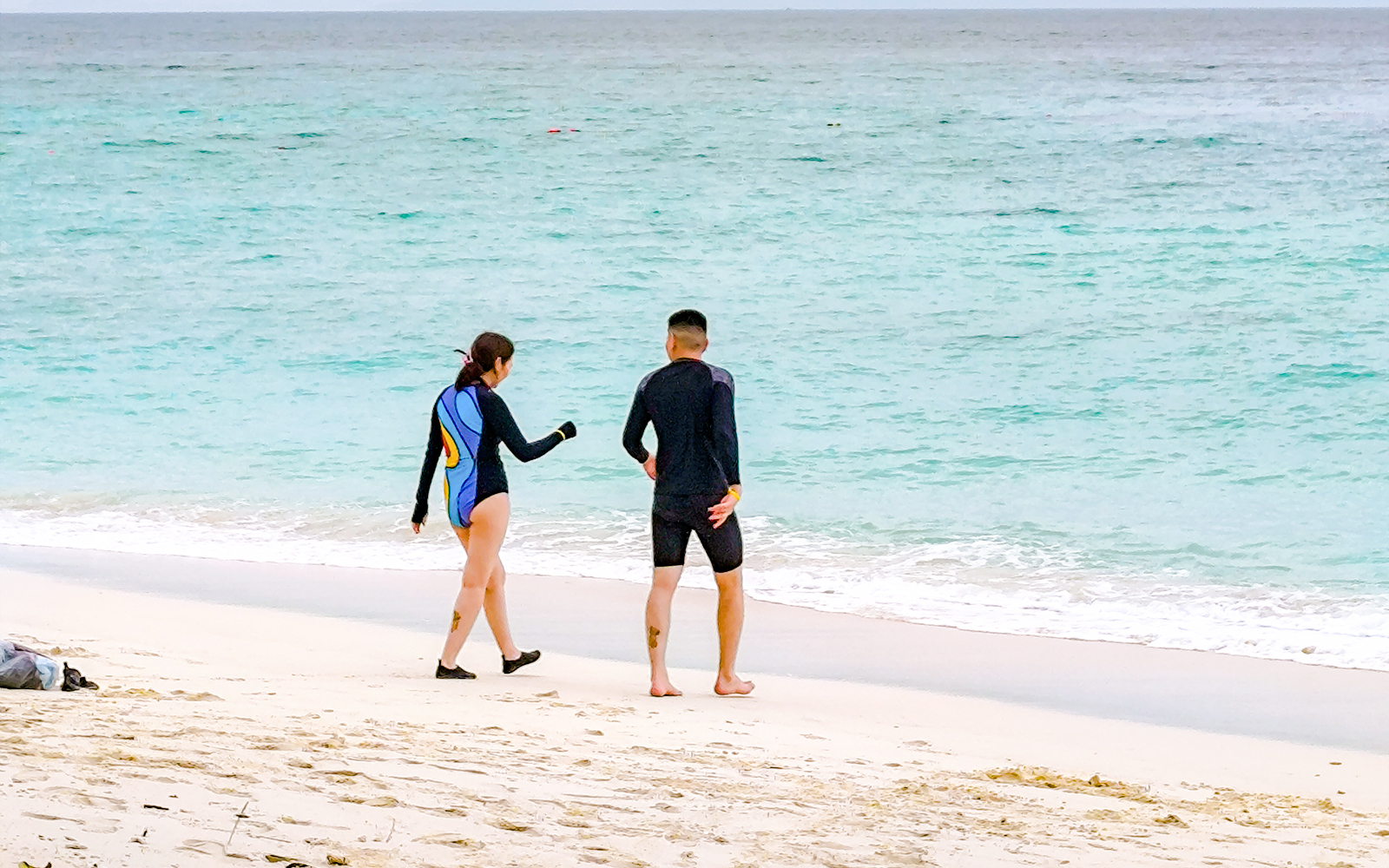 Couple in wetsuits walking on a white sand beach towards the ocean with distant islands.