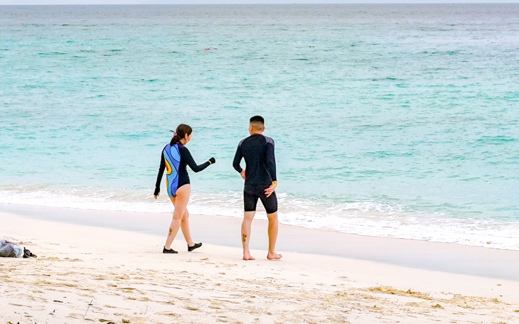 Couple in wetsuits walking on a white sand beach towards the ocean with distant islands.