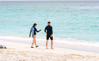 Couple in wetsuits walking on a white sand beach towards the ocean with distant islands.