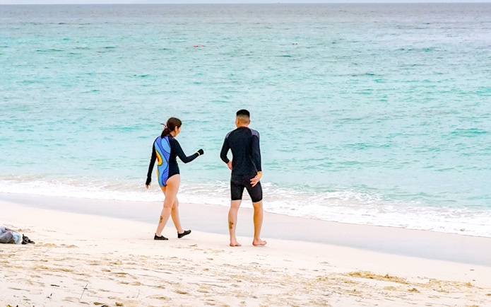 Couple in wetsuits walking on a white sand beach towards the ocean with distant islands.
