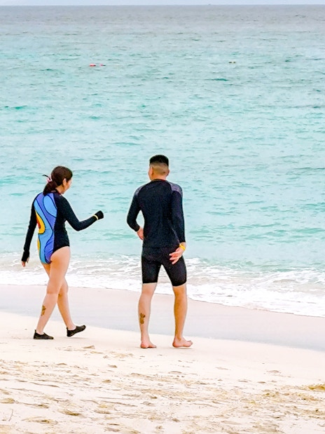 Couple in wetsuits walking on a white sand beach towards the ocean with distant islands.