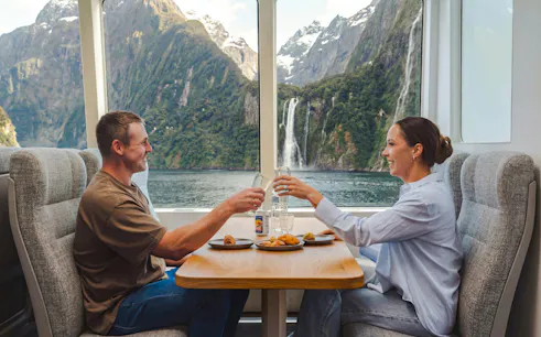 Couple enjoying a meal with a view of Milford Sound waterfall on a VIP cruise from Queenstown.