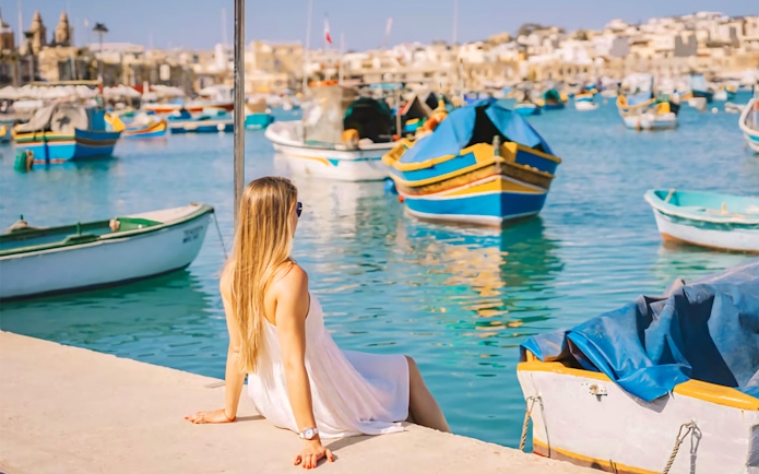 Woman sitting by colorful boats in Marsaxlokk Fishing Village, Malta.