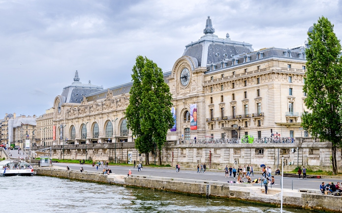 Musée d'Orsay exterior with people along the Seine River in Paris.