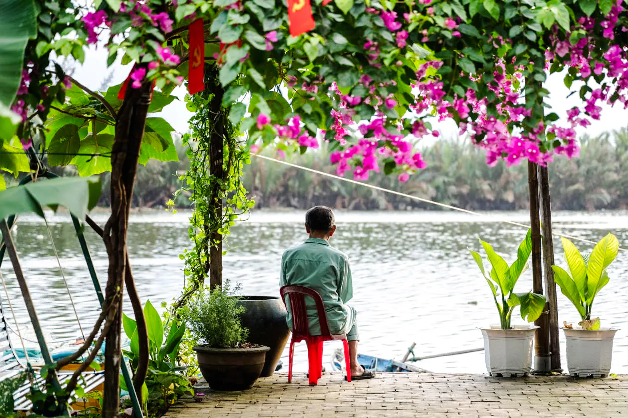 Man sitting by a river under flowering vines in Hoi An countryside.
