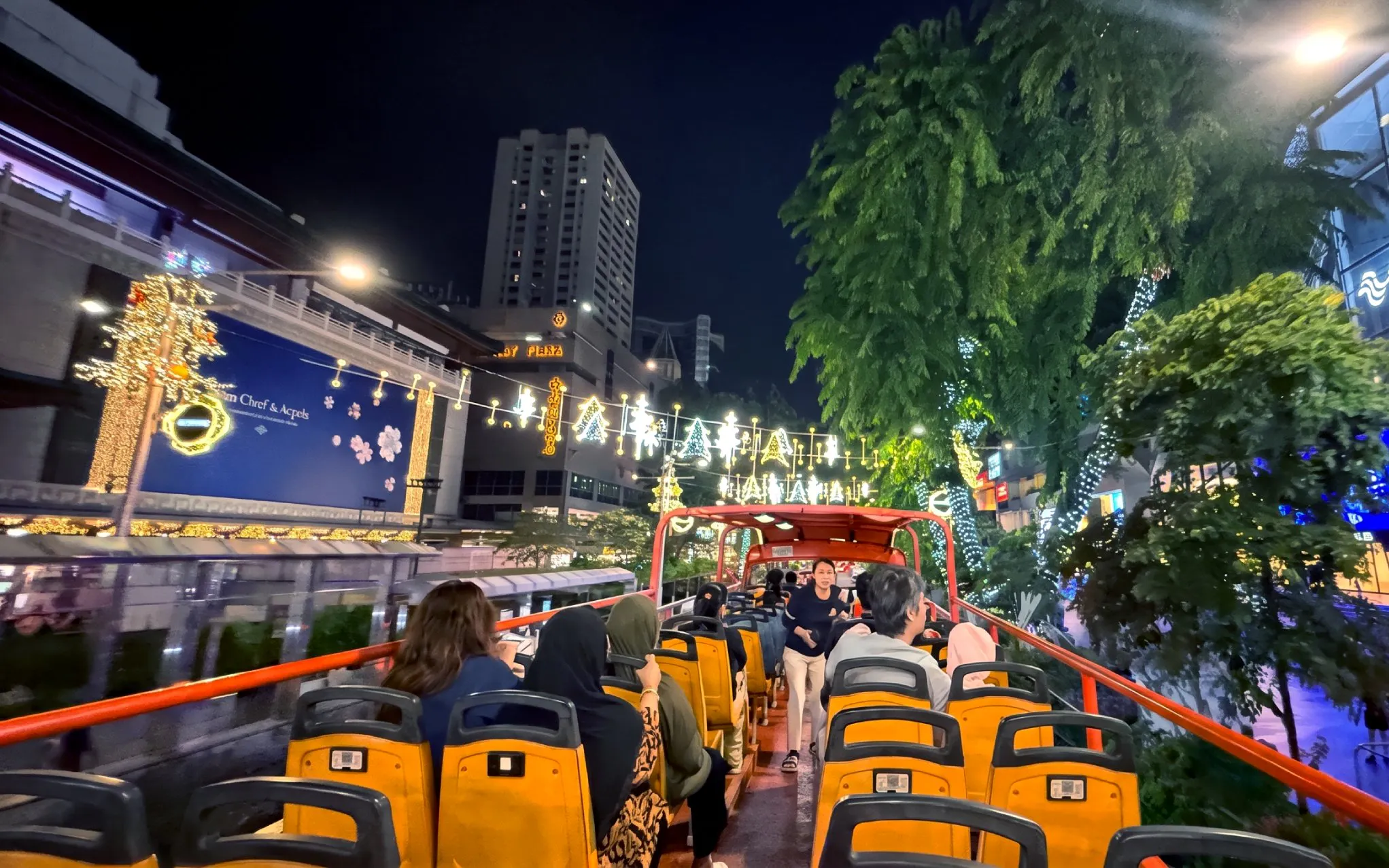 Passengers on an open-top double-decker bus at night under festive Christmas lights in a city street.