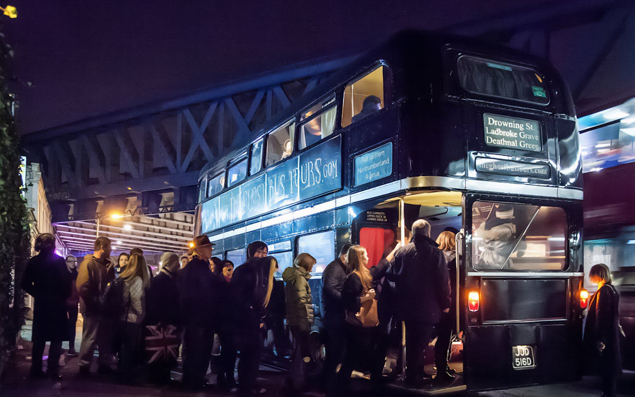 Nighttime crowd boarding a double-decker bus for a Ghost Bus Tour under a bridge.