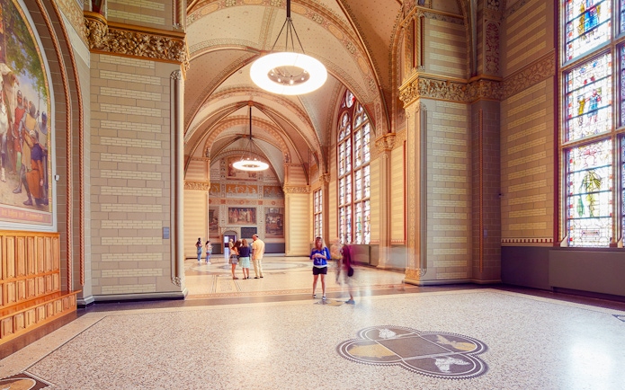 Visitors in The Great Hall at Rijksmuseum, Amsterdam, with stained glass windows and ornate ceilings.