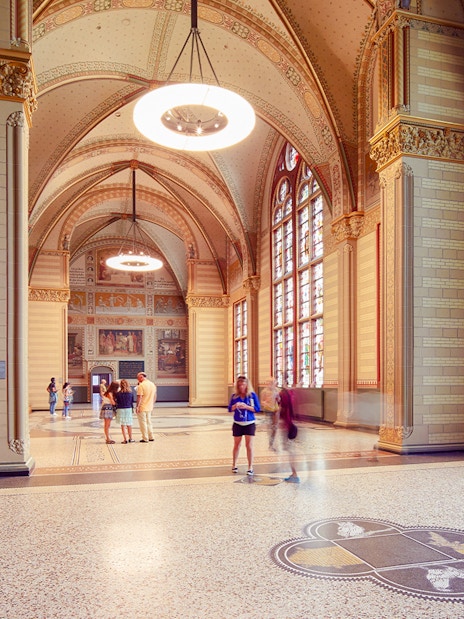 Visitors in The Great Hall at Rijksmuseum, Amsterdam, with stained glass windows and ornate ceilings.