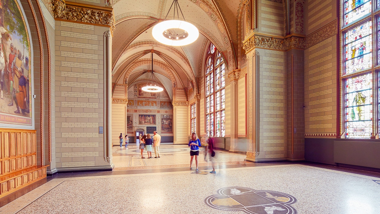Visitors in The Great Hall at Rijksmuseum, Amsterdam, with stained glass windows and ornate ceilings.