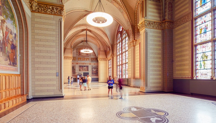Visitors in The Great Hall at Rijksmuseum, Amsterdam, with stained glass windows and ornate ceilings.