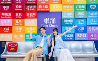 Couple sitting in front of colorful Tung Chung sign, preparing for Ngong Ping 360 cable car on Lantau Island.
