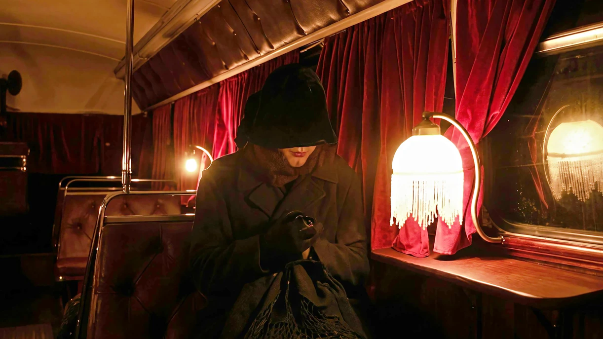 Ghost tour participant in dark cloak on vintage bus with red curtains and dim lighting.