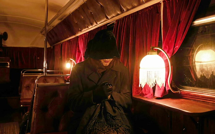 Ghost tour participant in dark cloak on vintage bus with red curtains and dim lighting.