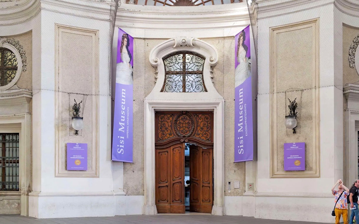 Entrance to the Sisi Museum with ornate wooden doors and purple banners in Vienna.