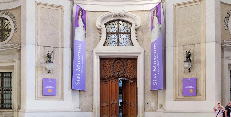 Entrance to the Sisi Museum with ornate wooden doors and purple banners in Vienna.