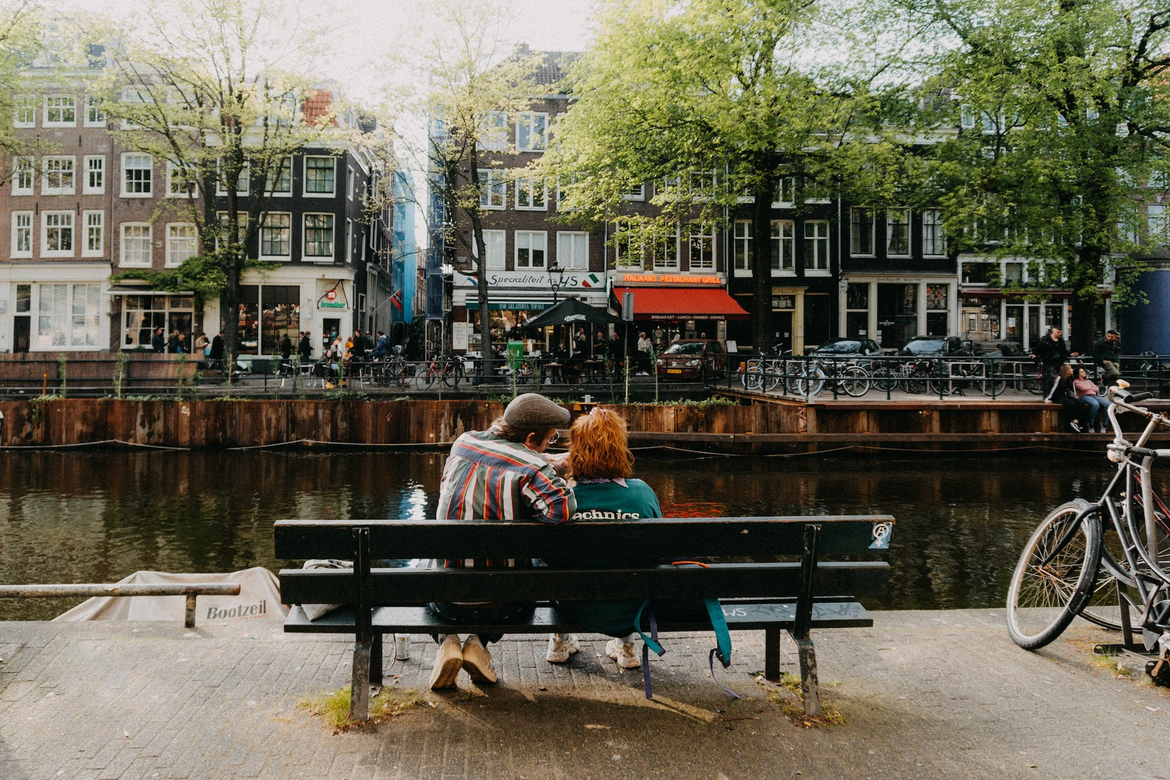 Couple sitting on a bench by a canal in Jordaan, Amsterdam, with bicycles and historic buildings nearby.