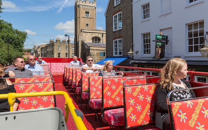Open-top bus with tourists on Cambridge Hop-On Hop-Off Tour passing historic buildings.