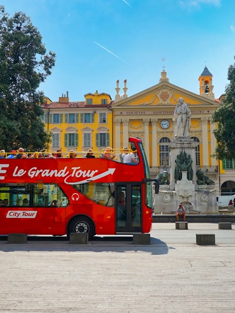 Hop-on hop-off bus in Nice, France, near a historic building and statue.