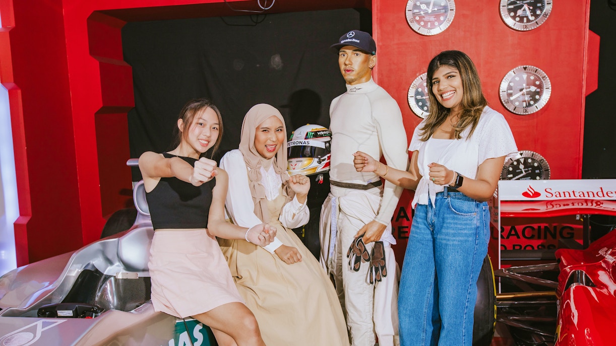 Visitors posing with Lewis Hamilton wax figure at Madame Tussauds London, near a race car display.