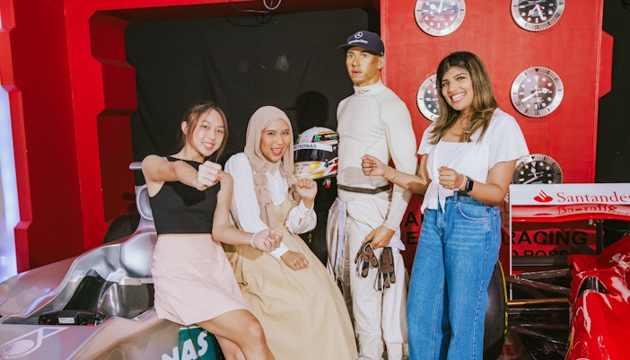 Visitors posing with Lewis Hamilton wax figure at Madame Tussauds London, near a race car display.
