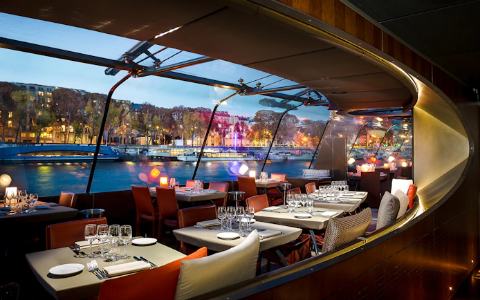 Dining area on a Seine River cruise boat with city view through large windows.