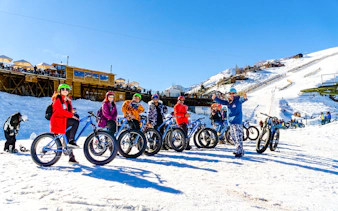 Group biking on snow at Farellones Park, Santiago, with ski slopes and lodge in background.