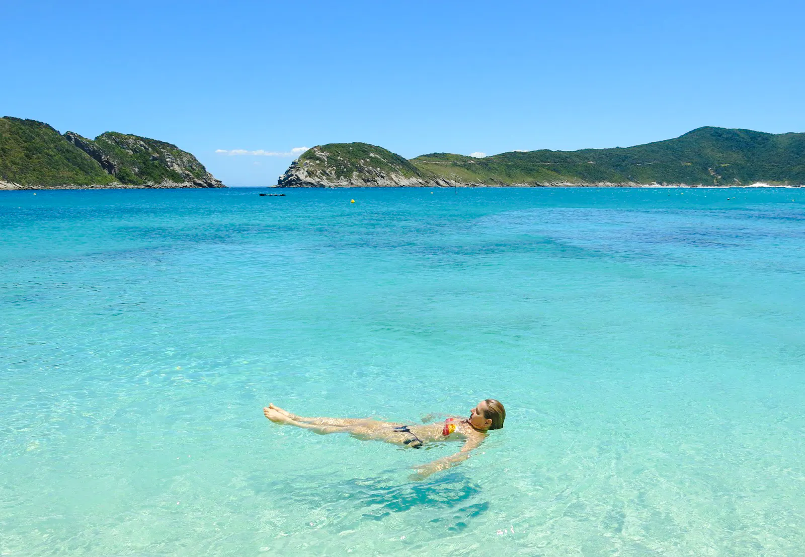 Person floating in clear turquoise water at Arraial do Cabo, Brazil, with green hills in the background.