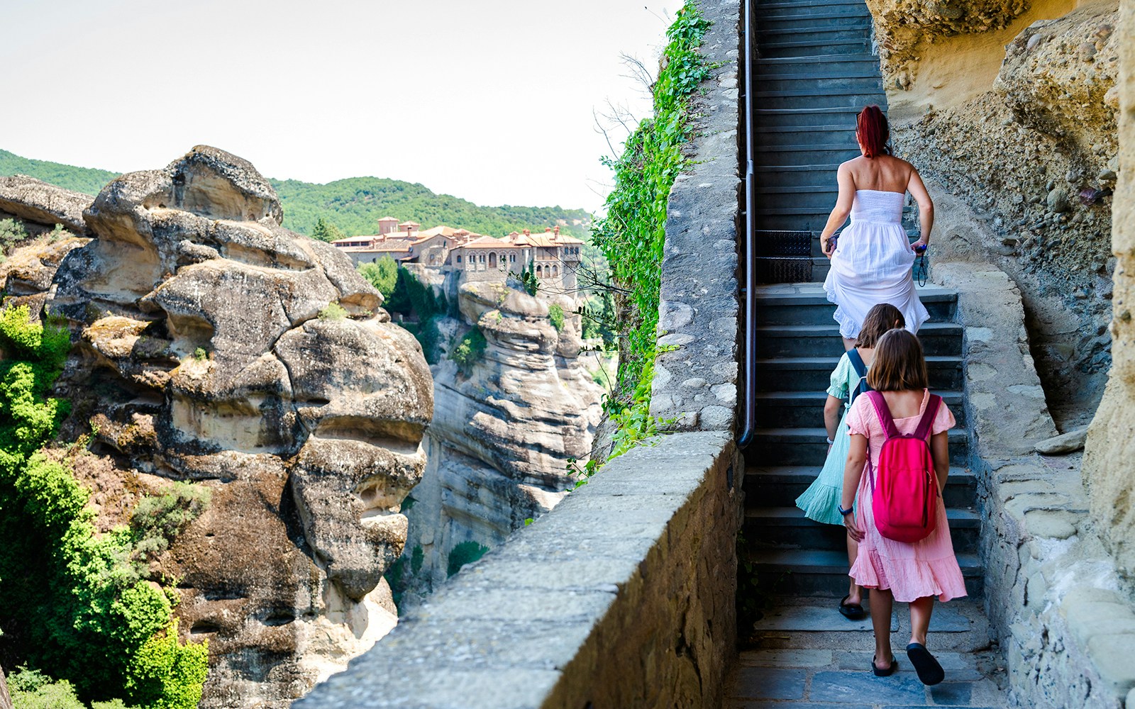 Family climbing stairs to Meteora monastery, Greece, with rock formations in view.