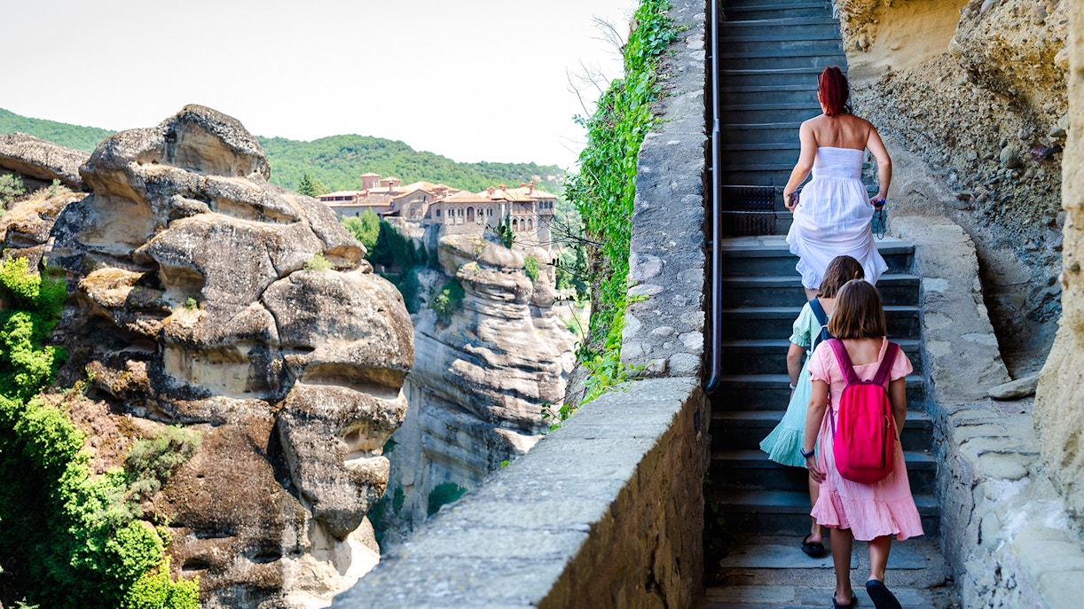 Family climbing stairs to Meteora monastery, Greece, with rock formations in view.