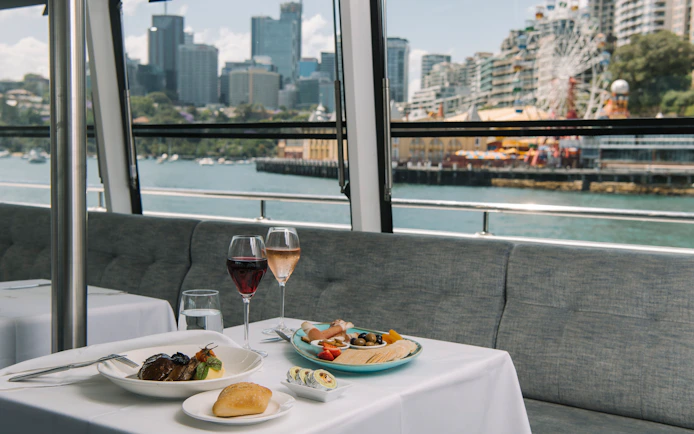 Dinner table with regional dishes and wine on a Sydney Harbour cruise, city skyline in the background.